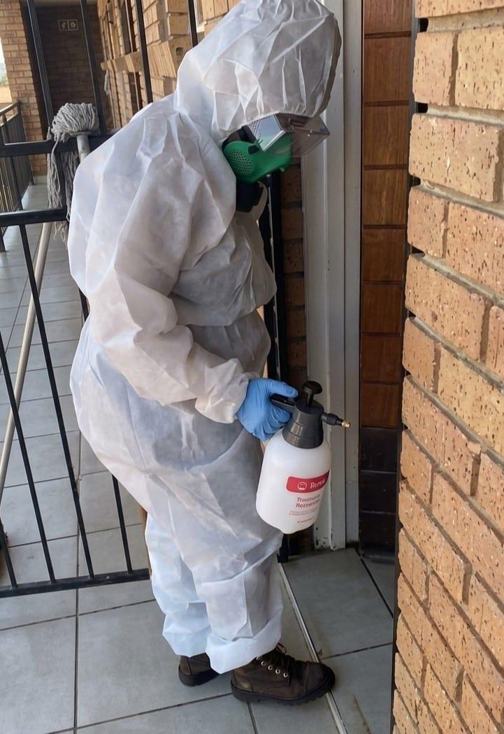 Beekay pest control technician in a protective coverall, respirator and gloves applying a targeted treatment along the brick wall of a residential building